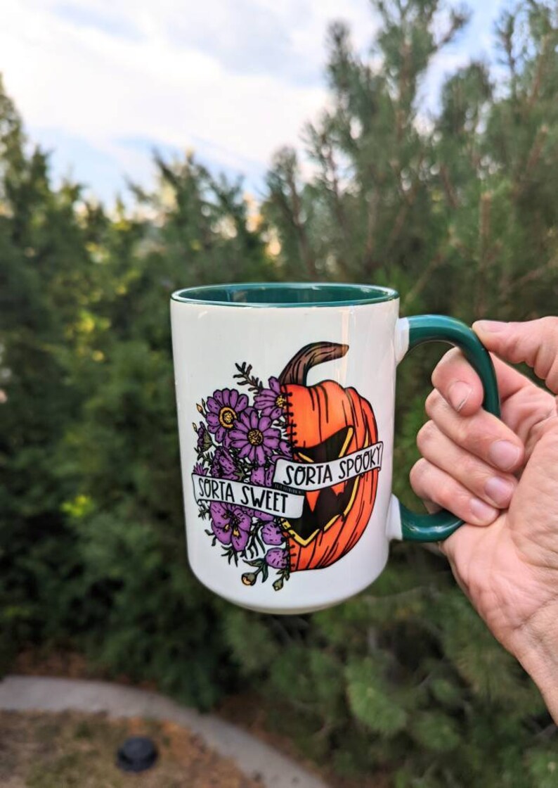May include: White ceramic mug with a green rim and handle. The mug features a graphic of a pumpkin with a stitched-up face, half of which is a jack-o'-lantern. The pumpkin is surrounded by purple flowers and a banner that reads "Sorta Sweet" on one side and "Sorta Spooky" on the other.