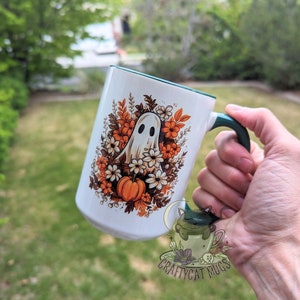 May include: White ceramic mug with a green rim and handle. The mug features a ghost surrounded by orange and white flowers and a pumpkin. The text "Craftycat Mugs" is printed on the bottom of the mug.
