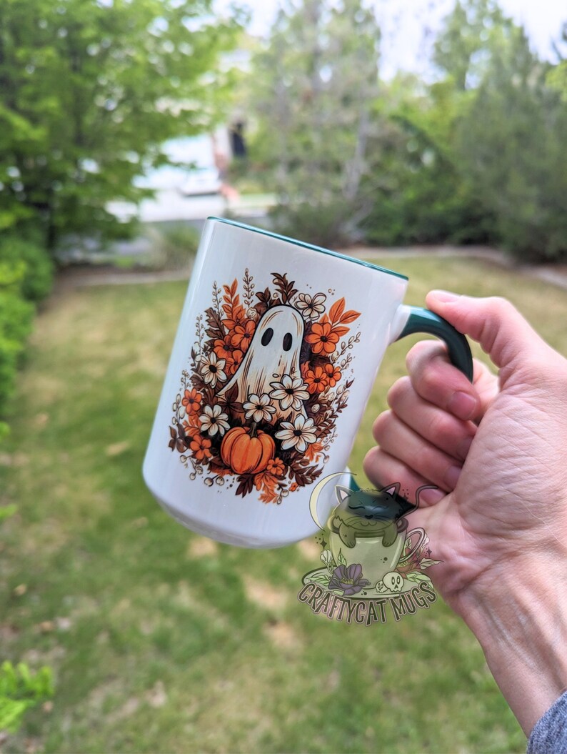 May include: White ceramic mug with a green rim and handle. The mug features a ghost surrounded by orange and white flowers and a pumpkin. The text "Craftycat Mugs" is printed on the bottom of the mug.