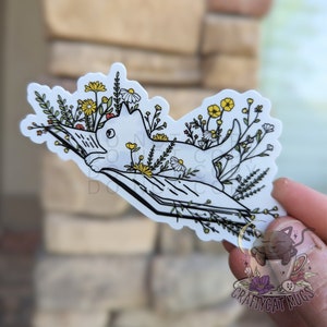 Op de afbeelding: Een witte kat die op een open boek ligt met bloemen die eromheen groeien. De kat kijkt naar links. Het boek is open en de pagina's zijn zichtbaar. De bloemen zijn in tinten geel, wit en roze.