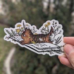Op de afbeelding: Een bruin-zwarte tabbykat die een boek leest, omringd door witte en gele bloemen. De kat ligt op zijn zij met zijn hoofd schuin, kijkend naar het boek. Het boek is open en heeft witte pagina's met zwarte lijnen. De bloemen zijn allemaal in verschillende vormen en maten en zijn rond de kat en het boek gerangschikt.