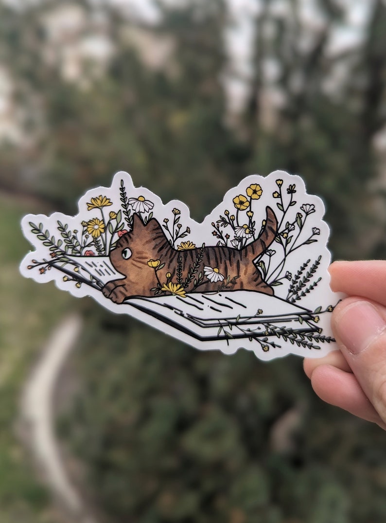 Op de afbeelding: Een bruin-zwarte tabbykat die een boek leest, omringd door witte en gele bloemen. De kat ligt op zijn zij met zijn hoofd schuin, kijkend naar het boek. Het boek is open en heeft witte pagina's met zwarte lijnen. De bloemen zijn allemaal in verschillende vormen en maten en zijn rond de kat en het boek gerangschikt.