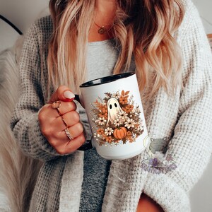 May include: White ceramic mug with a black rim and handle. The mug features a ghost with a pumpkin and fall flowers in orange, brown, and white.