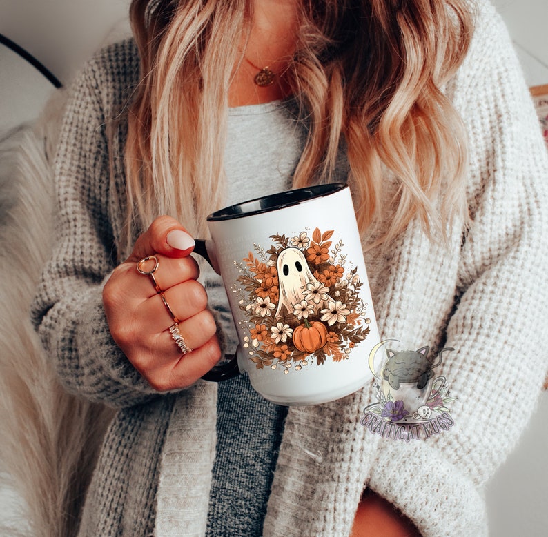 May include: White ceramic mug with a black rim and handle. The mug features a ghost with a pumpkin and fall flowers in orange, brown, and white.