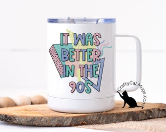 May include: White travel mug with a black silhouette of a cat and the text "It was better in the 90s" in a colorful, geometric design.