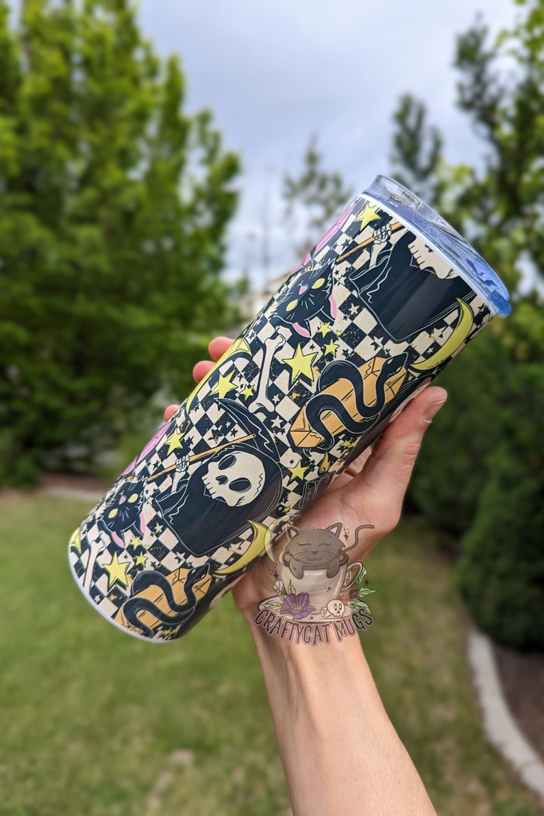 May include: A black and white tumbler with a checkered pattern and a graphic of a cat, a snake, and a skeleton wearing a hooded robe. The tumbler has a blue lid and is held by a hand. The text "CRAFTYCAT MUGS" is visible on the hand.