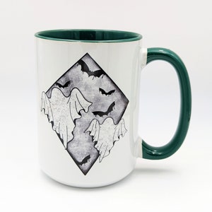 May include: White ceramic mug with a green rim and handle. The mug features a black and white illustration of ghosts and bats in a diamond shape.