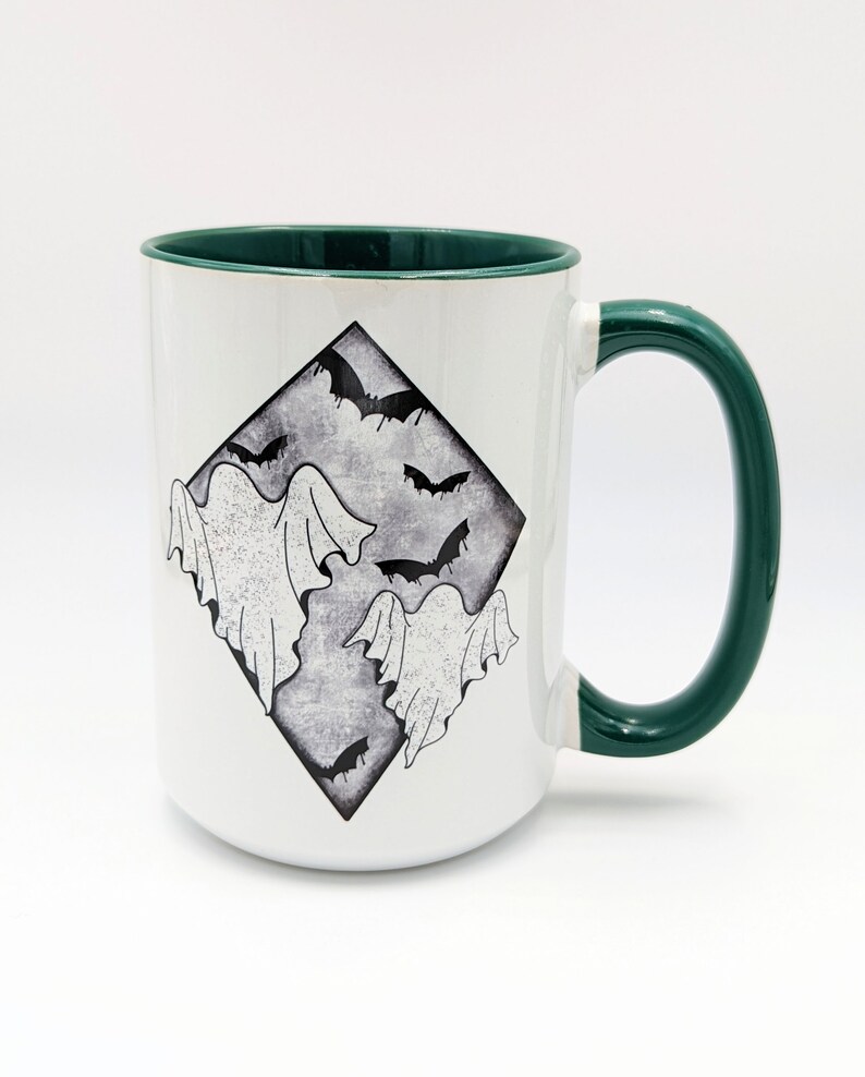 May include: White ceramic mug with a green rim and handle. The mug features a black and white illustration of ghosts and bats in a diamond shape.
