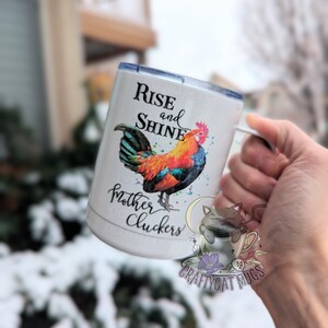 May include: White stainless steel travel mug with a colorful rooster illustration and the text "Rise and Shine Mother Cluckers". The mug has a black cat illustration with a coffee cup and the text "CraftyCat Mugs".