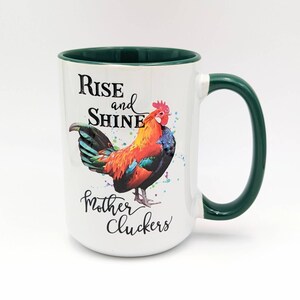 May include: White ceramic mug with a green rim and handle. The mug features a colorful rooster illustration and the text "Rise and Shine Mother Cluckers".
