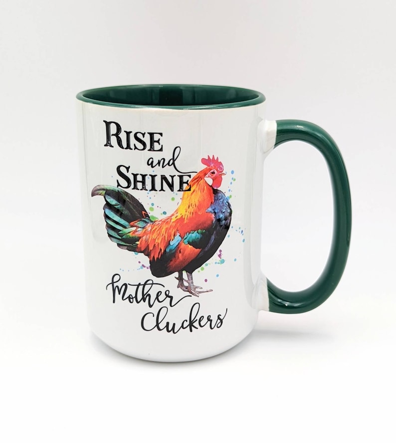 May include: White ceramic mug with a green rim and handle. The mug features a colorful rooster illustration and the text "Rise and Shine Mother Cluckers".