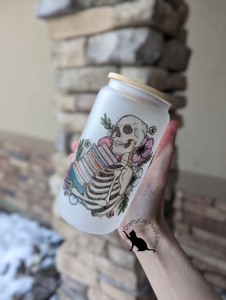 May include: A frosted glass water bottle with a bamboo lid features a hand-drawn design of a skeleton holding a stack of books. The design includes flowers and butterflies in pink, purple, and green. The bottle has the text "CraftyCatMugs.com" on the side.