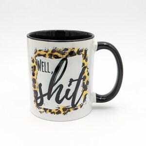 May include: White ceramic coffee mug with a black rim and handle. The mug features a leopard print design with a black square frame and the text "Well, shit" in black.