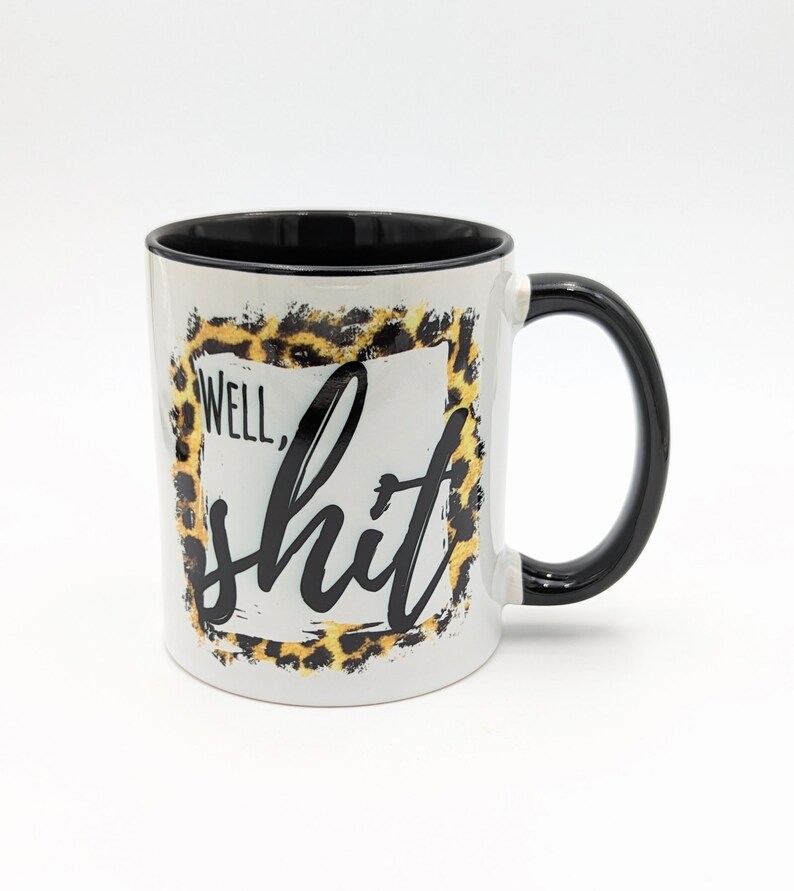 May include: White ceramic coffee mug with a black rim and handle. The mug features a leopard print design with a black square frame and the text "Well, shit" in black.