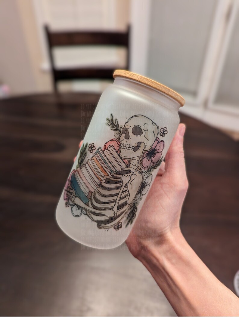 May include: A frosted glass jar with a bamboo lid features a hand-drawn illustration of a skeleton holding a stack of books. The skeleton is surrounded by flowers and leaves. The jar is a unique and whimsical way to store items.