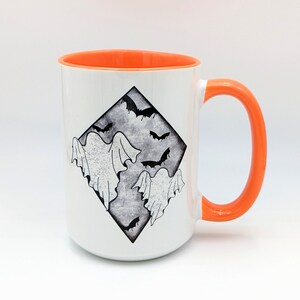 May include: White ceramic mug with an orange rim and handle. The mug features a black and white illustration of ghosts and bats in a diamond shape.