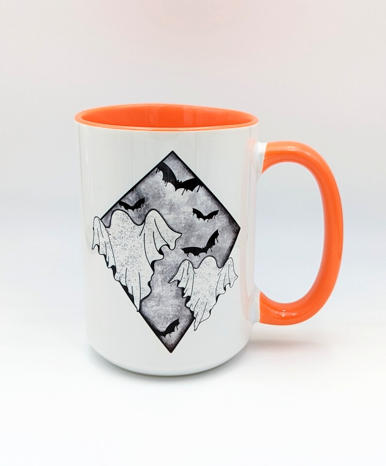 May include: White ceramic mug with an orange rim and handle. The mug features a black and white illustration of ghosts and bats in a diamond shape.