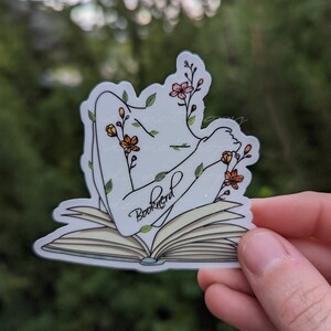 May include: A white sticker with a line drawing of a person with their arms outstretched, surrounded by flowers and leaves, lying on an open book. The book has the word "Booknerd" written on it.