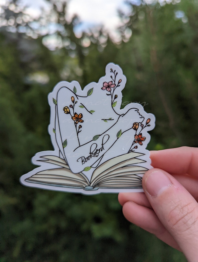 May include: A white sticker with a line drawing of a person with their arms outstretched, surrounded by flowers and leaves, lying on an open book. The book has the word "Booknerd" written on it.