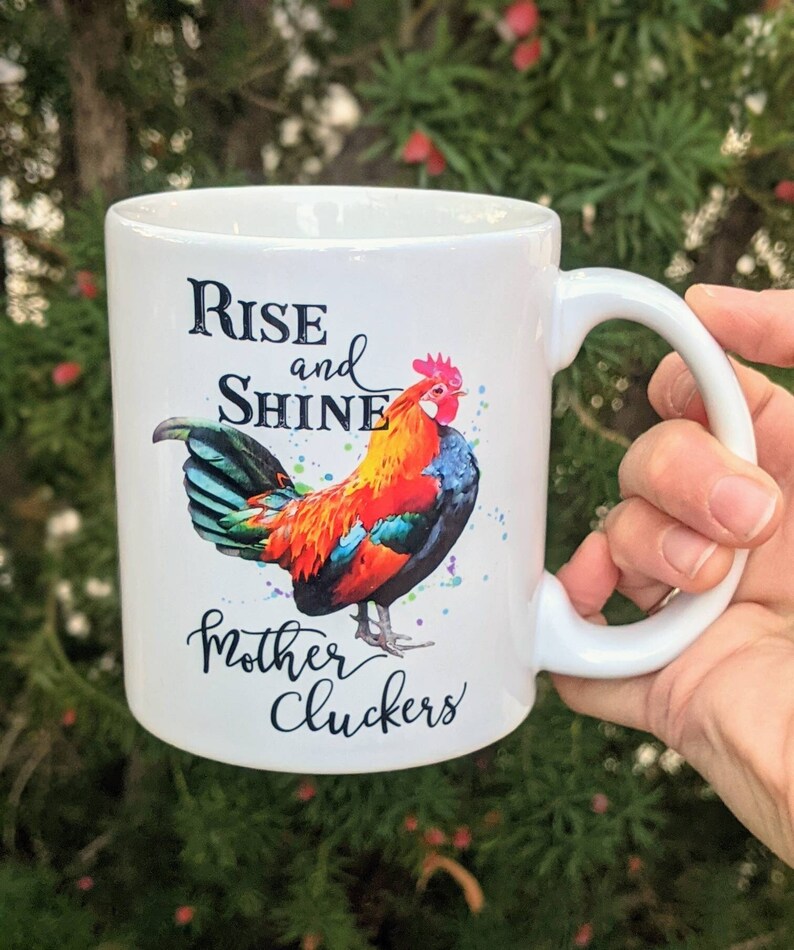 May include: White ceramic coffee mug with a colorful rooster illustration and the text "Rise and Shine Mother Cluckers".