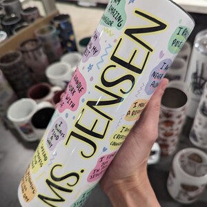 Op de afbeelding: Een witte tumbler met een zwarte en gele inscriptie die "Ms. Jensen" luidt. De tumbler is versierd met kleurrijke, handgeschreven affirmaties, waaronder "Ik ben geweldig", "Ik vind altijd een manier" en "Ik ben creatief".