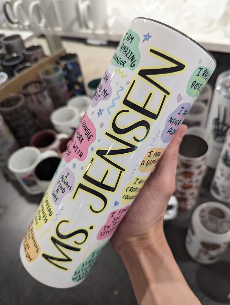 Op de afbeelding: Een witte tumbler met een zwarte en gele inscriptie die "Ms. Jensen" luidt. De tumbler is versierd met kleurrijke, handgeschreven affirmaties, waaronder "Ik ben geweldig", "Ik vind altijd een manier" en "Ik ben creatief".