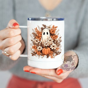 May include: White ceramic mug with a ghost and fall foliage design. The ghost is white with black eyes and is surrounded by orange, brown, and white flowers. The mug has a stainless steel lid.