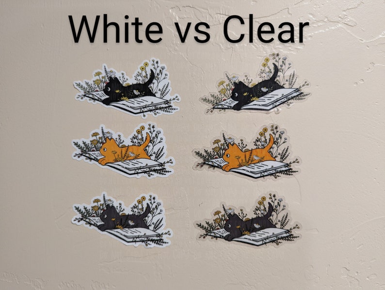 Op de afbeelding: Een vergelijking van twee katten stickers op een witte achtergrond. De stickers tonen een zwarte kat en een oranje kat die een boek lezen, omringd door bloemen. De tekst "White vs Clear" staat bovenaan de afbeelding.