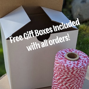 K&ouml;nnte beinhalten: Wei&szlig;e Geschenkschachtel aus Pappe mit dem Text "Free Gift Boxes Included with all orders!" auf der Vorderseite. Die Schachtel ist ge&ouml;ffnet und eine Spule roter und wei&szlig;er B&auml;ckergarn befindet sich im Vordergrund.