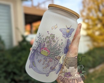 May include: A frosted glass tumbler with a bamboo lid, decorated with a whimsical design. The illustration includes a cauldron with a frog, mushrooms, and plants, along with a moth and other botanical elements. The words "Craftycat Mugs" are also visible.