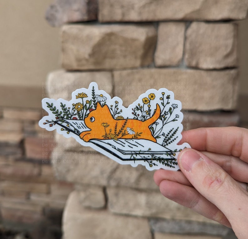 Op de afbeelding: Een oranje kat die op een boek ligt met een witte achtergrond. De kat wordt omringd door kleine gele en witte bloemen.