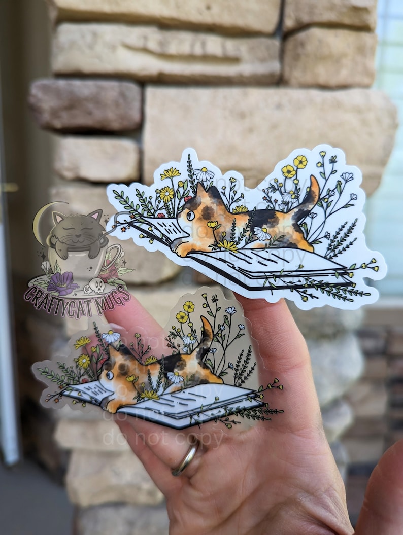 Op de afbeelding: Twee transparante stickers met een driekleurige kat die een boek leest, omringd door bloemen. De kat ligt op het boek in de ene sticker en zit op het boek in de andere sticker. De stickers bevinden zich op een hand.