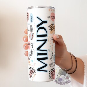 May include: White stainless steel tumbler with a black and blue design. The tumbler has the name "MINDY" written in large black letters. The tumbler is decorated with a variety of abstract shapes and patterns in pink, blue, and black.