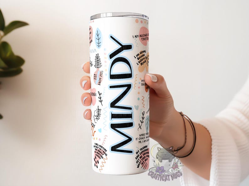 May include: White stainless steel tumbler with a black and blue design. The tumbler has the name "MINDY" written in large black letters. The tumbler is decorated with a variety of abstract shapes and patterns in pink, blue, and black.