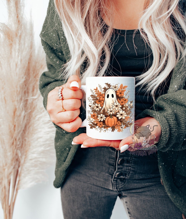 May include: White ceramic mug with a ghost and fall foliage design. The ghost is white and has a surprised expression. The foliage is in shades of orange, brown, and yellow. There are also small white flowers and a pumpkin.