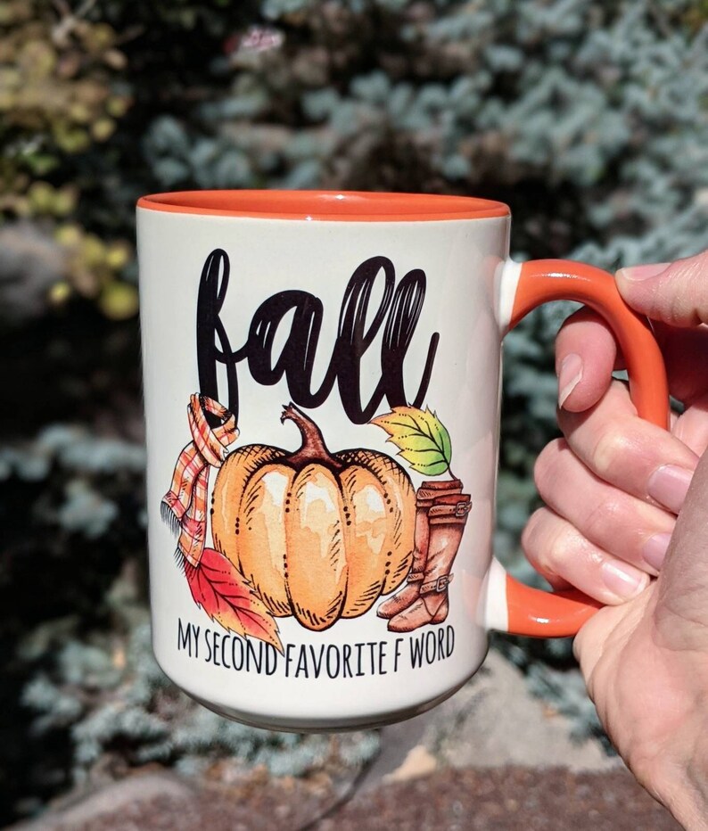 May include: White ceramic mug with an orange rim and handle. The mug features a black and white illustration of a pumpkin with fall leaves and boots. The text "fall" is written in black cursive and the text "my second favorite f word" is written in black below the pumpkin.