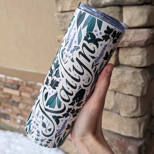 Puede incluir: Un vaso de acero inoxidable blanco con un dise&ntilde;o floral verde y morado y el nombre "Caylyn" escrito en cursiva negra.