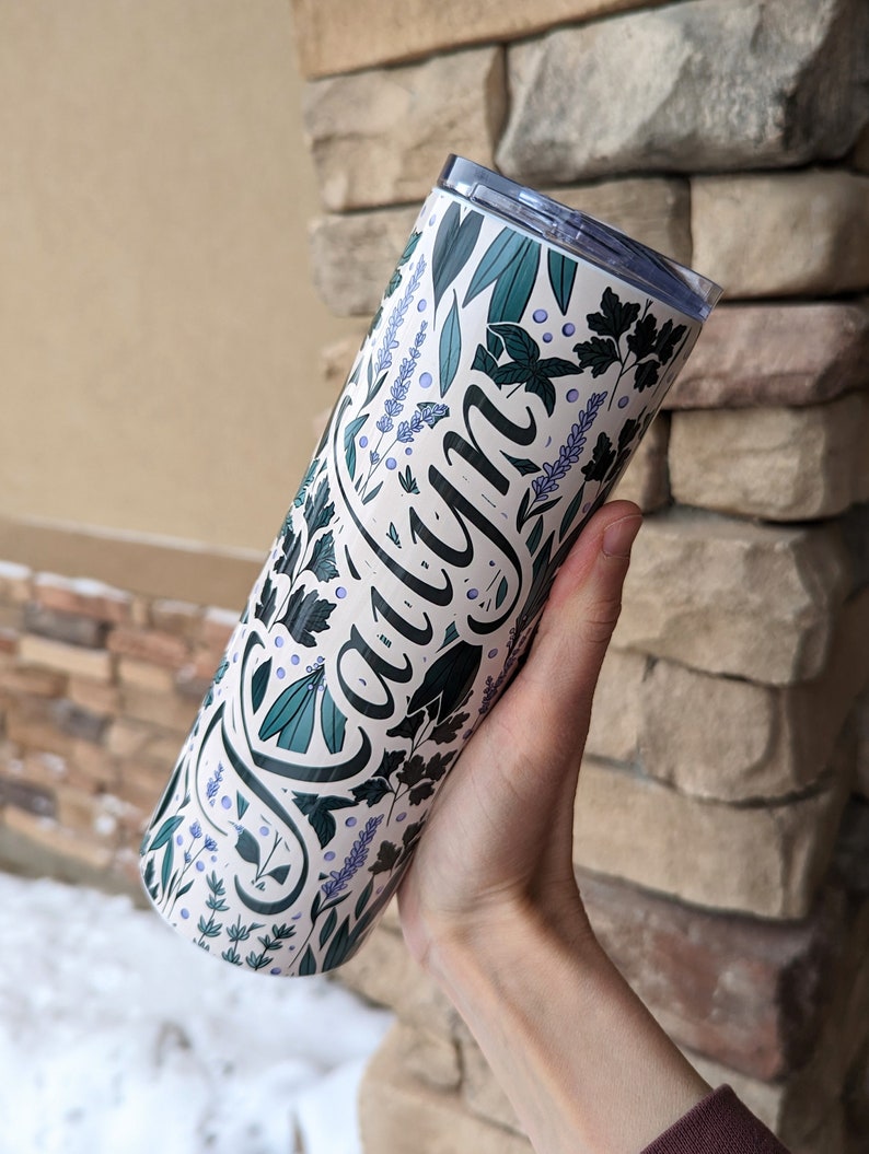 Puede incluir: Un vaso de acero inoxidable blanco con un dise&ntilde;o floral verde y morado y el nombre "Caylyn" escrito en cursiva negra.