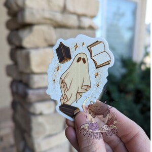 May include: A white ghost with a book in its hand, surrounded by books and sparkles. The ghost is standing on a book. The image is on a white background with a black and white cat in a teacup in the bottom right corner. The text "CRAFTYCAT MUGS" is below the cat.