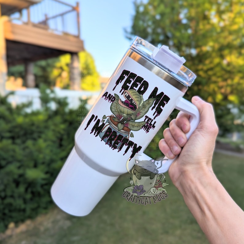 May include: White stainless steel travel mug with a black and green graphic of a plant with a mouth and the text "Feed Me and Tell Me I'm Pretty".