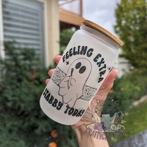 May include: A frosted glass tumbler with a bamboo lid. The tumbler features a cartoon ghost design with the text "Feeling Extra Stabby Today!". The design is in black and white. The tumbler is held in front of a blurred outdoor background.