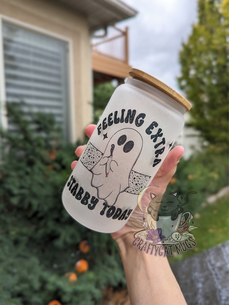 May include: A frosted glass tumbler with a bamboo lid. The tumbler features a cartoon ghost design with the text "Feeling Extra Stabby Today!". The design is in black and white. The tumbler is held in front of a blurred outdoor background.