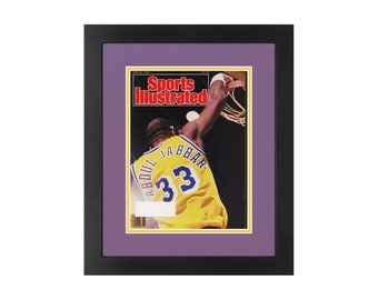 May include: A framed Sports Illustrated magazine cover featuring a basketball player in mid-air, wearing a yellow jersey with the number 33 and the name "Abdul-Jabbar" on the back. The cover is dated June 24, 1985.