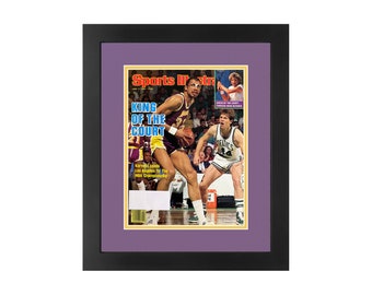 May include: A framed Sports Illustrated magazine cover featuring Kareem Abdul-Jabbar in a purple and gold Lakers uniform, dribbling the ball, with a Boston Celtics player guarding him. The cover headline reads "King of the Court".