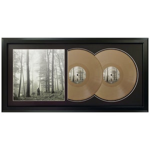 May include: A black framed double album display featuring a black and white photograph of a forest with a person walking in the distance. The two vinyl records are gold in colour.