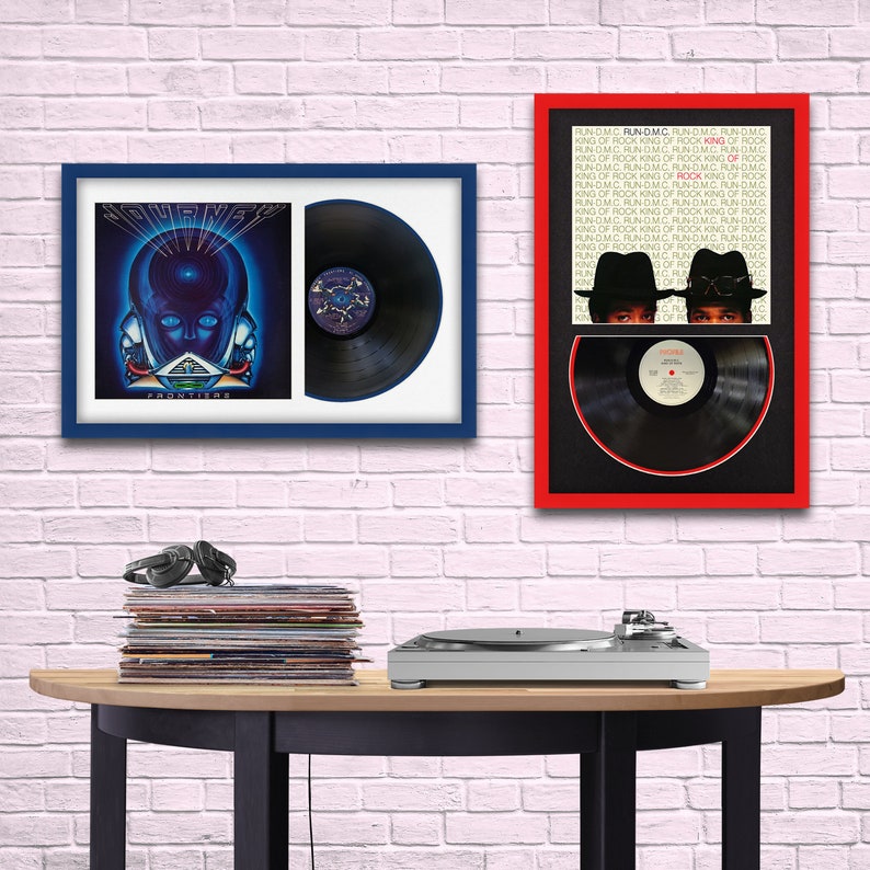 May include: Two framed vinyl records, one with a Journey album cover and the other with a Run-DMC album cover. Both frames are red and blue. A turntable and a stack of vinyl records are on a wooden table.