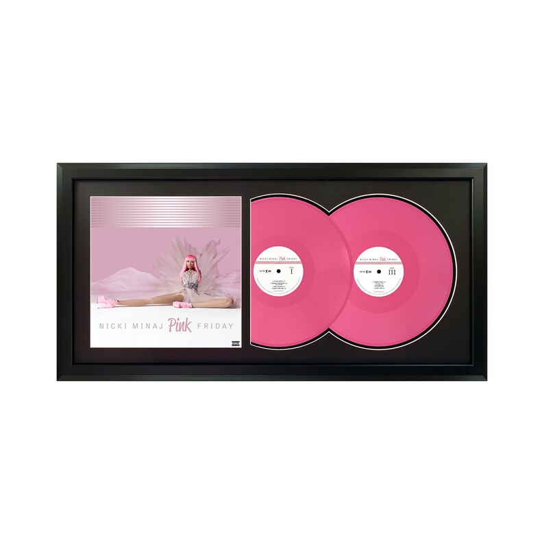 May include: A framed double vinyl record set of Nicki Minaj's Pink Friday album. The album cover features Nicki Minaj in a pink wig and white outfit, sitting on a pink cloud. The vinyl records are pink and have the album title printed on them.