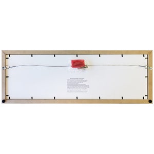 May include: The back of a rectangular wooden picture frame with a wire hanging system and a small paper with assembly instructions. The frame is light brown and has black metal corners.