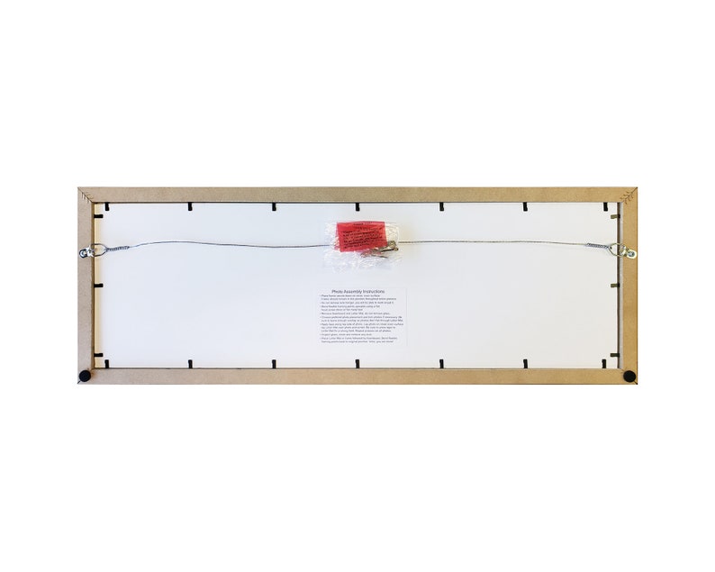 May include: The back of a rectangular wooden picture frame with a wire hanging system and a small paper with assembly instructions. The frame is light brown and has black metal corners.