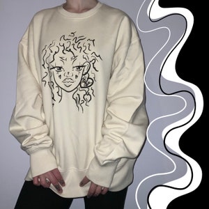 Puede incluir: Una sudadera de color crema con una ilustración en blanco y negro de la cara de una mujer con un corazón en la oreja. La ilustración está estilizada con líneas onduladas.